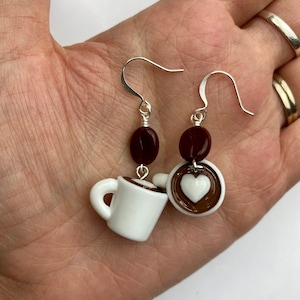 May include: A pair of coffee-themed earrings. Each earring features a white mug with a brown rim, a dark brown coffee bean, and a coffee cup with a white heart design. The earrings have silver-colored hooks.