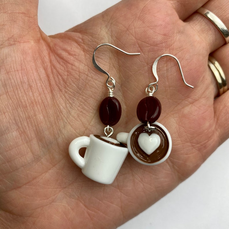 May include: A pair of coffee-themed earrings. Each earring features a white mug with a brown rim, a dark brown coffee bean, and a coffee cup with a white heart design. The earrings have silver-colored hooks.