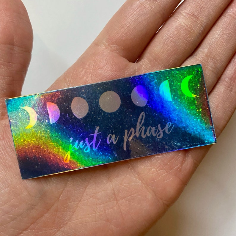 May include: A holographic sticker with a rainbow iridescent background and a black silhouette of the moon in different phases. The text "just a phase" is written in white below the moon phases.