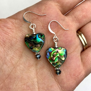 Puede incluir: Un par de pendientes de plata en forma de corazón con detalles de concha de abulón iridiscente y cristal negro.