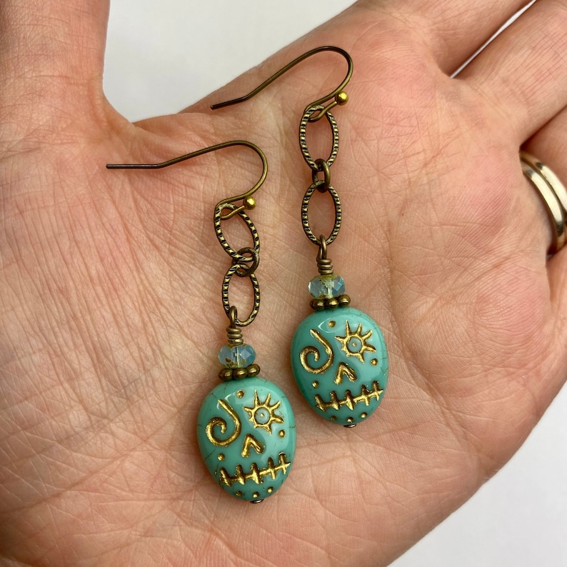 May include: A pair of turquoise blue skull earrings with gold accents. The earrings are made with a chain and a skull-shaped bead.