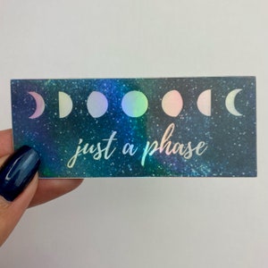 May include: A holographic sticker with a galaxy background and a row of iridescent moons. The text "just a phase" is written in white below the moons.