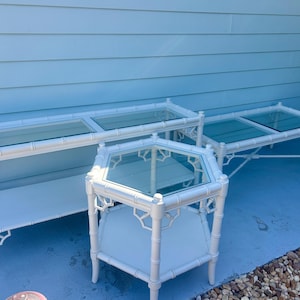 May include: A set of three white tables featuring a faux bamboo frame design. The collection includes one long rectangular console table, one rectangular coffee table, and one hexagonal side table, each topped with clear glass panels.