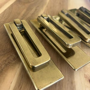 May include: Close-up of several brass drawer pulls. Each pull has a rectangular base with a rectangular handle. The pulls are arranged on a wooden surface. The brass has a warm, golden hue.