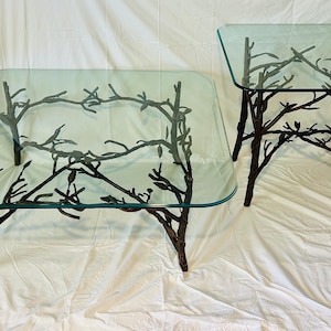 May include: A set of two tables featuring clear square glass tops supported by dark brown metal bases designed to resemble intertwined tree branches with small leaves. The larger table is rectangular, while the smaller table is square.