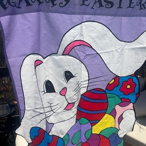 May include: A purple Easter flag with the words "HAPPY EASTER" in black. The flag features a white bunny with pink accents and colorful Easter eggs. The eggs are decorated with various patterns and colors.