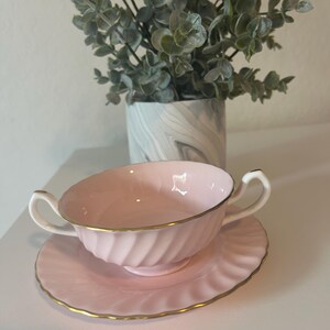 May include: A pink teacup and saucer set with a gold trim. The cup has a swirl design and the saucer has a scalloped edge.