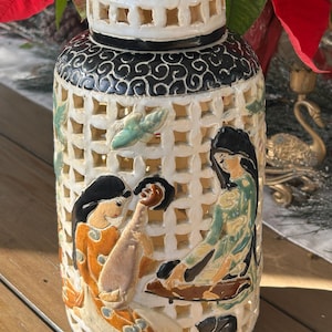 May include: A decorative ceramic jar with a lid. The jar has a lattice design with painted figures in orange, green, and black. The lid is white with a black swirl pattern. The jar is a decorative item.