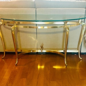 May include: A semi-circular console table featuring a clear glass top supported by a polished gold-tone metal frame. The base consists of four curved legs connected by a horizontal stretcher bar with a central decorative finial detail.