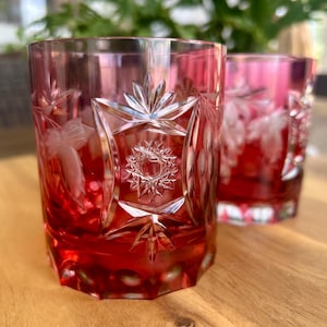 May include: Two ruby red glass tumblers featuring intricate cut glass patterns. Each vessel displays a prominent starburst motif surrounded by etched leaf designs and a faceted base, showcasing a deep crimson hue and reflective glass surfaces.