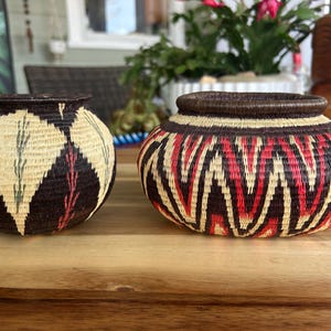 May include: Two hand-woven baskets. The basket on the left has a dark brown background with cream-coloured diamond shapes and red and green accents. The basket on the right has a red, cream, and dark brown zigzag pattern.