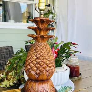 Puede incluir: Una lámpara de mesa de madera con forma de piña con un soporte de lámpara de metal dorado. La lámpara tiene un diseño tallado y una bombilla. La lámpara está sobre una mesa de madera.
