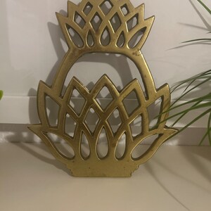 May include: A gold-toned metal trivet featuring a stylised pineapple shape with an openwork lattice design. The object has a flat base and a pointed top, constructed from a solid, reflective material with a smooth, uniform metallic finish.