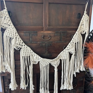 May include: A cream-colored macrame wall hanging featuring a series of interconnected triangular knotted patterns. The piece is suspended by two metal hooks and includes long, dangling fringe sections that hang vertically from the bottom edge.