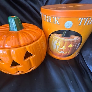 May include: A ceramic orange pumpkin-shaped jar with a green stem lid and triangular cutouts for eyes and a mouth, positioned next to an orange metal cylindrical container featuring the text TRICK OR TREAT and a graphic of the pumpkin jar.