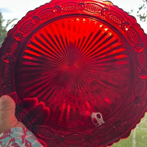 May include: A circular ruby red glass plate featuring a central sunburst pattern of radiating lines. The outer rim is decorated with a repeating embossed geometric motif consisting of oval shapes and cross-hatched textures along the scalloped edge.