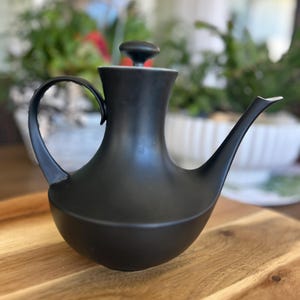 May include: A black ceramic teapot with a curved handle and a long spout. The teapot has a rounded base and a small, round lid. It is displayed on a wooden surface.