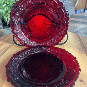 Könnte beinhalten: Zwei runde Teller aus rubinrotem Glas mit gewelltem Rand und einem detaillierten Pressmuster. Das Design umfasst ein zentrales Strahlenmotiv, das von einer komplexen Bordüre aus geprägten Ovalen und schraffierten Texturen am Rand umgeben ist.