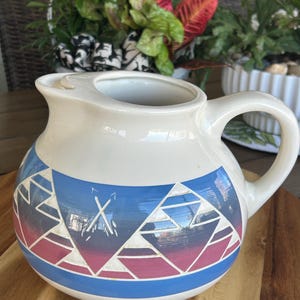 May include: Cream ceramic pitcher with a rounded body and a handle. The pitcher has a geometric design in blue, pink, and white around the middle. The design includes triangles and crossed arrows. The pitcher is on a wooden surface.