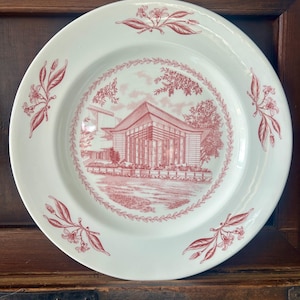May include: A white ceramic plate featuring a red transferware design. The center depicts a modern building with large windows surrounded by a leafy border. Four floral sprigs are evenly spaced around the wide rim of the plate.