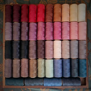 Peut inclure: Un plateau en bois rempli de bobines de fil de différentes couleurs. Les couleurs des fils vont des rouges et violets foncés aux roses clairs, bleus et verts. Les bobines sont soigneusement disposées, présentant un large éventail de teintes.