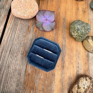 Puede incluir: Un joyero de terciopelo azul marino con tres ranuras, rodeado de varias piedras y una flor de hortensia púrpura sobre una superficie de madera. La caja está abierta, lista para guardar anillos.