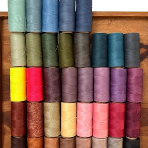 Peut inclure: Plusieurs bobines de fil colorées, disposées en rangées. Les bobines sont de différentes nuances, notamment bleu, vert, jaune, rouge et marron. Le texte "Linhasita 0,35" est en haut de l'image.
