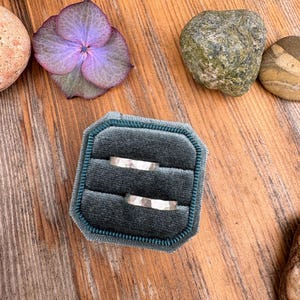 Puede incluir: Dos anillos de plata con superficies texturizadas descansan en un joyero de terciopelo verde azulado. El joyero está sobre una superficie de madera, rodeado de piedras decorativas y una flor de hortensia morada. Anillos de boda o compromiso.
