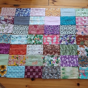 May include: A selection of folded fabric face coverings in diverse colours and patterns, including floral, paisley, and geometric designs. The masks are arranged on a wooden surface, showcasing a variety of styles and prints.