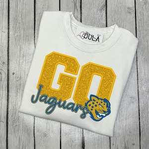 Puede incluir: Sudadera blanca con el texto "GO Jaguars" en purpurina dorada y letras azules. Una mascota de jaguar azul y dorado está en la parte inferior derecha. La sudadera tiene cuello redondo y una etiqueta que dice "OU LA Customs".