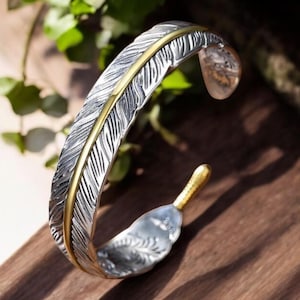 Puede incluir: Brazalete de puño plateado con diseño de pluma y detalles dorados. El brazalete es abierto y curvado, con un grabado detallado de pluma. El acento dorado recorre el centro de la pluma.