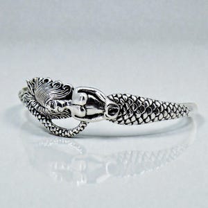 May include: A silver mermaid bracelet. The cuff bracelet features a detailed mermaid design with flowing hair, a serene face, and a scaled tail. The mermaid figure is the central decorative element of the bracelet.