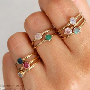 Puede incluir: Se muestran varios anillos dorados, cada uno con una pequeña gema de colores. Las gemas incluyen rosa, verde, azul y rojo. El tamaño de la piedra es de aproximadamente 4 mm.