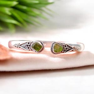 May include: A silver bangle bracelet with two green gemstone accents. The bracelet has a delicate, ornate design.