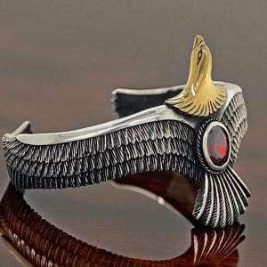Peut inclure: Bracelet manchette argenté avec un motif d'aigle. Le bracelet présente une tête d'aigle dorée et une pierre rouge ovale. Les ailes sont finement détaillées avec des motifs de plumes. Un bijou élégant.