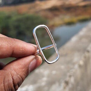 May include: A silver ring with a rectangular shape and a horizontal bar across the centre.