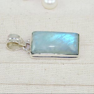 May include: A rectangular pendant with a light blue, iridescent gemstone set in a silver-colored metal frame. The pendant has a loop at the top for a chain. The gemstone has a subtle, shimmering effect, with hints of blue and white.