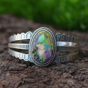 May include: Silver cuff bracelet with an oval, iridescent gemstone in green, purple, and cream. The bracelet has a rope-like border around the stone and an openwork design on the band. The bracelet is approximately 15 cm in length.