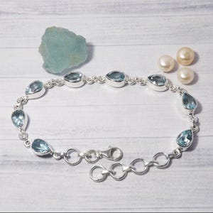 May include: A silver chain bracelet with teardrop-shaped blue topaz gemstones. The bracelet has a lobster clasp closure and an extender chain.