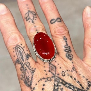May include: Oval-shaped, red gemstone ring with a silver-colored, ornate setting. The ring's gemstone is a vibrant, glossy red. The ring is worn on a finger with multiple tattoos.