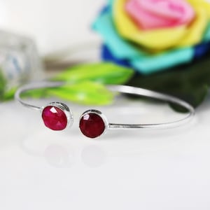 Puede incluir: Una pulsera abierta de plata con dos piedras preciosas de rubí rojo oscuro, redondas y facetadas, engastadas en cada extremo. La banda de metal es fina y pulida, creando un marco circular sencillo que resalta el color de las piedras.