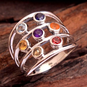 May include: A silver ring with a unique design, featuring seven colorful gemstones. The gemstones are set in a row, with a variety of colors including blue, purple, orange, and red. The ring is displayed on a wooden surface.