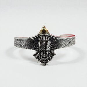 May include: Silver cuff bracelet with an eagle motif. The eagle's head is gold-coloured, and the wings are outstretched. This bracelet is a striking accessory, perfect for adding a touch of nature-inspired style to any outfit.
