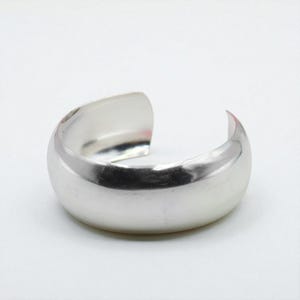 Puede incluir: Un brazalete de plata pulida. El diseño abierto presenta una superficie redondeada y lisa. El brazalete tiene un acabado brillante y reflectante, sobre un fondo blanco. Esta joya es un accesorio sencillo pero elegante.