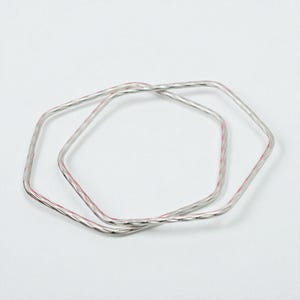 May include: Two silver-colored, square-shaped bangles with a subtle pink hue running through the center. The bangles are intertwined, set against a plain white background. The design is simple and modern.