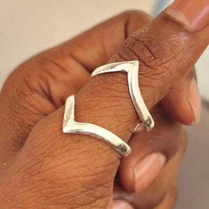 Puede incluir: Un anillo de pulgar de plata con un diseño en forma de doble V. El anillo se lleva en el pulgar de una persona.