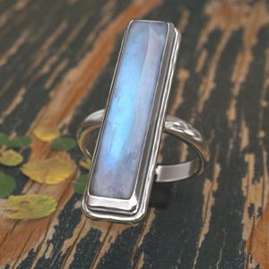 Puede incluir: Un anillo de plata con una piedra de luna rectangular engastada en un engaste de bisel. El anillo está sobre una superficie de madera.