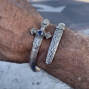 Puede incluir: Un brazalete de plata con forma de espada que presenta un pomo con cabeza de león, una piedra preciosa circular azul en la guarda y un emblema de escudo con un león. El brazalete incluye una pieza de plata separada con un intrincado nudo celta.