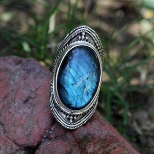 May include: A silver ring with a large oval blue labradorite gemstone. The ring has a detailed design with a beaded border.