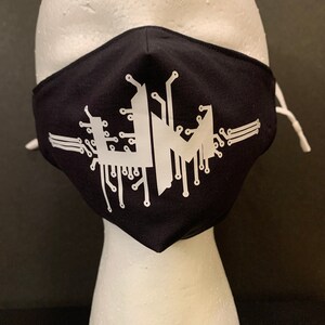 Puede incluir: Máscara facial negra con un diseño de circuito impreso blanco. El diseño presenta una letra "M" estilizada con líneas y conectores de circuito impreso.